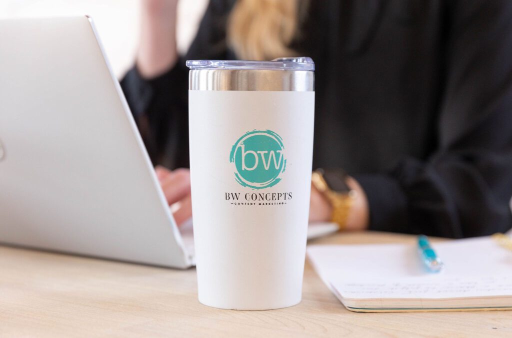 BW Concepts branded tumbler focus on wooden desk with laptop, notebook, pen, and person in background. 